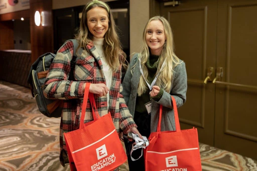 Two young women clad in business-casual attire proudly standing and smiling with red InsightsEDU-branded tote bags in hand.