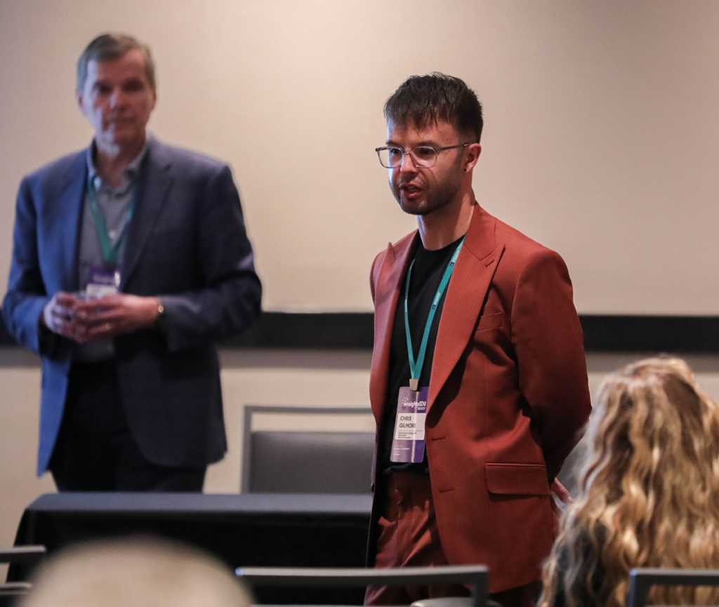 Dr. Chris Gilmore is dressed in a burnt orange blazer speaking on multi-channel engagement strategies to craft a seamless student experience. Dr. Tom Green from Salesforce stands behind a table offering additional insights.
