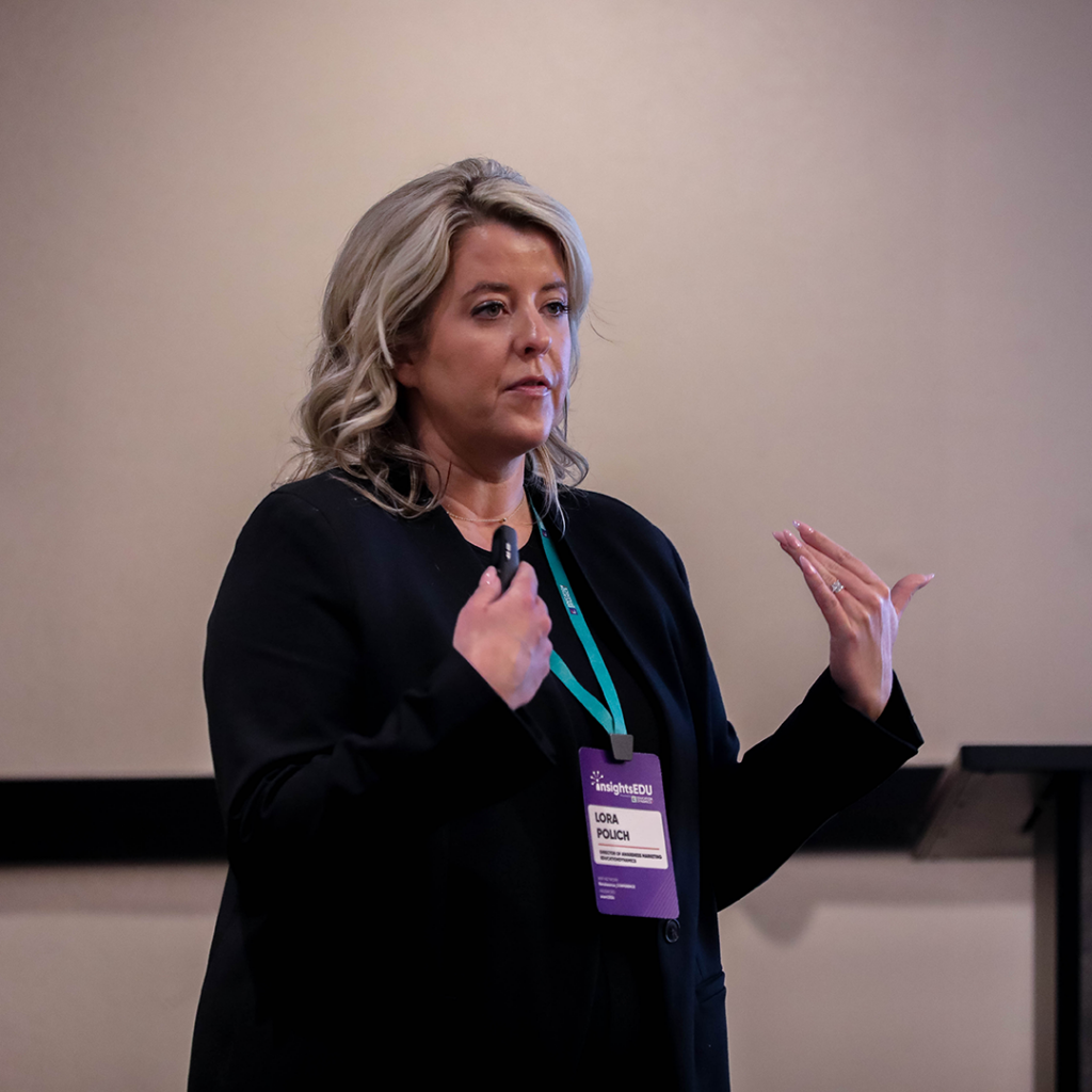 Lora Polich, Director of Awareness Marketing at EducationDynamics is speaking to an audience who is off camera. She is gesturing to herself as she holds a presentation remote control