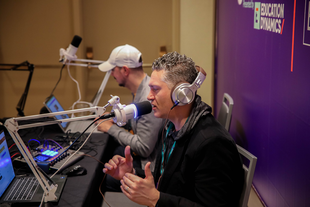 Joe Sallustio is sitting at a recording station recording a live session of  The EdUp Experience Podcast at InsightsEDU. They are in front of a EducationDynamics wall art. Co-founder Elvin Freytes is also at the recording table monitoring sounds on a laptop. 