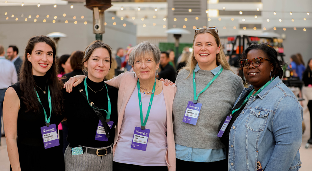 InsightsEDU attendees are grouped together on the Skyline Terrace in Phoenix, AZ. They have their arms over each other's shoulders and have market lights, a header and other conference attendees behind them.